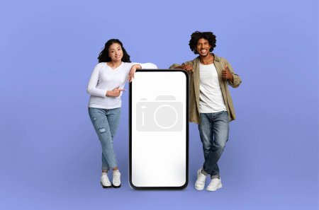 Foto de Un hombre y una mujer afroamericanos alegres posan junto a una maqueta de teléfonos inteligentes de gran tamaño sobre un fondo sólido - Imagen libre de derechos