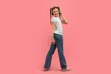 Young caucasian teen girl strolls with a joyful attitude wearing headphones, isolated on a pink background, presenting a carefree youngster vibe