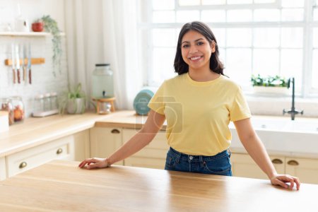 Une femme arabe joyeuse avec un sourire éclatant se tient avec confiance dans sa cuisine maison, présentant une atmosphère chaleureuse et accueillante