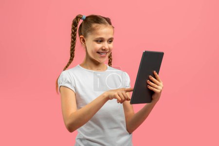 Téléchargez les photos : Une jeune fille joyeuse aux cheveux tressés interagit avec une tablette sur un fond rose vif, regardant engagé - en image libre de droit