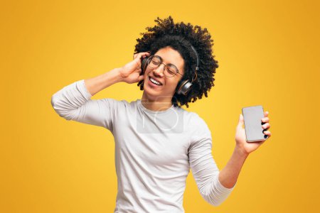 Photo for Handsome young african-american guy in wireless headphones listening to music online on smartphone and dancing, orange background - Royalty Free Image