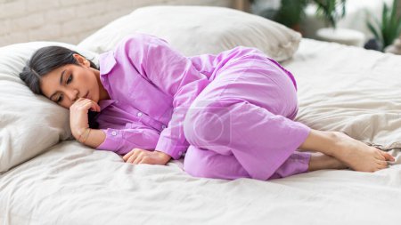 Frustrated unhappy woman in a purple shirt is lying on a white bed with a blurred face, suggesting depression and anxiety