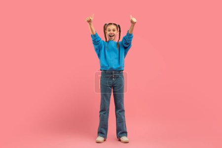 Téléchargez les photos : Une jeune fille joyeuse dans un pull bleu et un jean debout avec les bras levés, donnant pouces vers le haut sur un fond rose - en image libre de droit