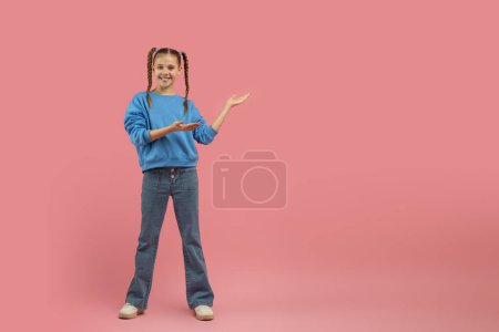 Photo for A cheerful young girl with braids in a blue sweater presenting or showing something to her side on a pink background, copy space - Royalty Free Image