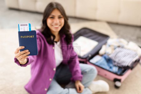 Photo for A smiling middle eastern woman sits on the floor showing her passport and boarding pass in front of an open suitcase, indicating travel preparation - Royalty Free Image
