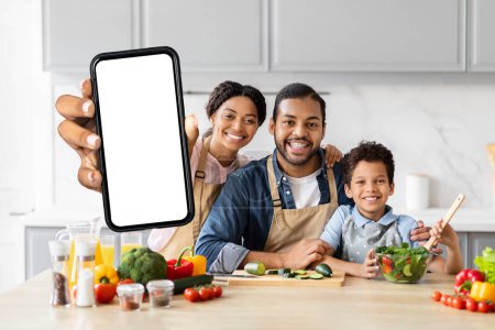 Foto de Familia afroamericana positiva cocinando juntos en la cocina, mostrando un teléfono inteligente con pantalla blanca en blanco, recomendando blog de alimentos - Imagen libre de derechos