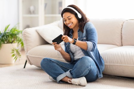 Foto de Mujer afroamericana está sentada en el suelo, interactuando con el dispositivo de teléfono inteligente, jugando juegos móviles - Imagen libre de derechos