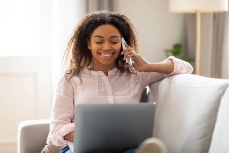 Téléchargez les photos : La femme afro-américaine sourit pendant qu'elle parle au téléphone et regarde son ordinateur portable, montrant un scénario multitâche à la maison - en image libre de droit