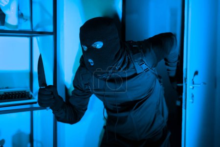 Ein maskierter Einbrecher in schwarzer Kleidung, der ein Messer in der Hand hält, wird bei einem nächtlichen Wohnungseinbruch auf frischer Tat ertappt, angestrahlt von gespenstischem Blaulicht