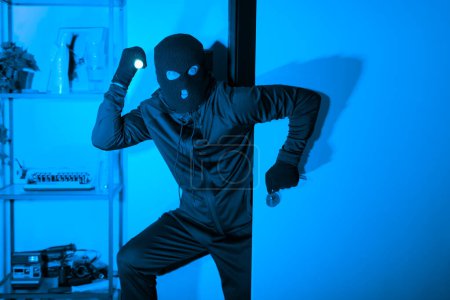 Téléchargez les photos : La silhouette d'un voleur pris en pleine action tenant une lampe de poche représente le vol, le danger et la vulnérabilité d'un appartement la nuit, capturé dans une intense teinte de bleu - en image libre de droit