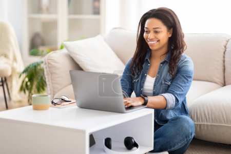 Femme afro-américaine occupée est assis sur le sol avec les jambes croisées, travaillant intensément sur un ordinateur portable, dactylographiant et cliquant. Il y a un sentiment de concentration et de productivité dans l'air.