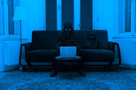 Téléchargez les photos : Un voleur dans un masque assis à une table, à l'aide d'un ordinateur portable, potentiellement voler des informations Scène dans un appartement, se sent étrange et dangereux la nuit - en image libre de droit
