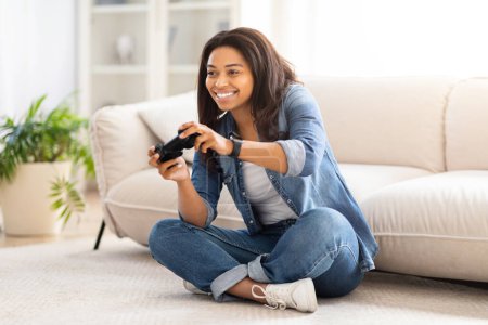 Téléchargez les photos : Femme afro-américaine assise sur le sol absorbée par un jeu vidéo. Elle tient un contrôleur, concentré sur l'écran. Autour d'elle sont des visuels de jeu colorés et des câbles se connectant à un - en image libre de droit
