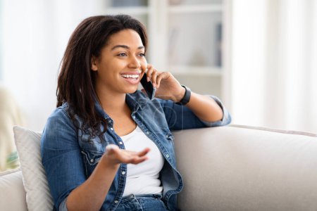 Téléchargez les photos : Une Afro-Américaine est assise sur un canapé, engagée dans une conversation téléphonique. Elle tient un téléphone portable à l'oreille avec une attention particulière, faisant des gestes occasionnels pendant l'appel. - en image libre de droit