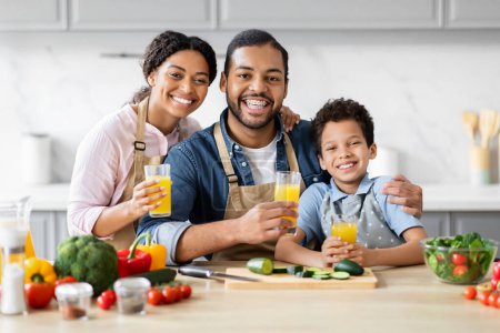 Foto de Padre afro-americano positivo madre e hijo bebiendo jugo de naranja fresco, familia feliz preparando ensalada de verduras saludables juntos en la cocina - Imagen libre de derechos