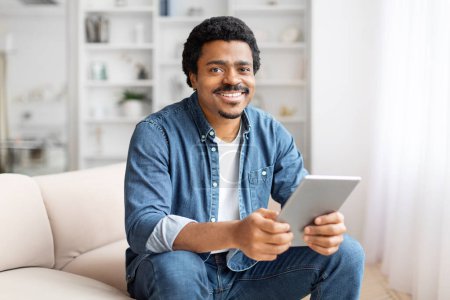 An engaging black man smiles while using his tablet at home, capturing the versatility of modern technology among people