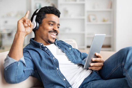 Photo for African american guy at home using a tablet with headphones on, indicating enjoyment of digital media - Royalty Free Image