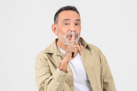 Photo for A quiet elderly man with a finger on his lips, isolated on white, encouraging silence, perhaps in a library or during a private moment - Royalty Free Image