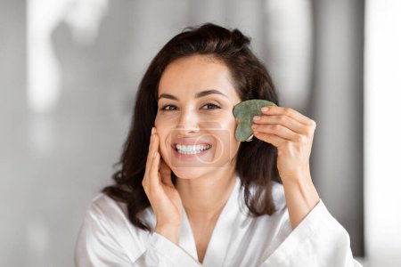 Une jeune femme joyeuse dans une chemise blanche tient un rouleau de massage facial vert contre sa joue, avec un accent sur les soins de la peau et la beauté