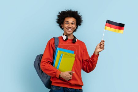 Téléchargez les photos : Étudiant afro-américain souriant aux cheveux touffus, posant avec le drapeau allemand et des livres colorés, représentant des études ou des voyages internationaux, sur un fond bleu isolé - en image libre de droit