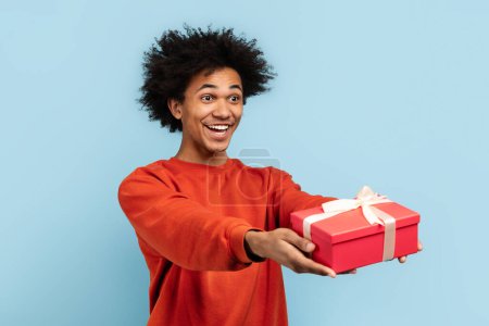 Photo for African american guy with a pleasant smile presenting a red gift box tied with a white ribbon, symbolizing generosity and festive occasions, against a blue isolated backdrop - Royalty Free Image