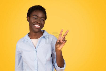 Foto de Con una sonrisa brillante, esta mujer afroamericana, un zoómetra de la generación z, hace un signo de paz, aislado en un amarillo - Imagen libre de derechos