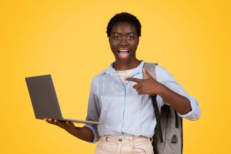 An excited african american woman from generation z, zoomer, points at her laptop screen, isolated against a yellow background