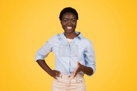 Eine selbstbewusste junge afrikanisch-amerikanische Frau, die mit den Händen auf den Hüften vor gelbem Hintergrund in die Kamera lächelt und Positivität und Zuversicht ausstrahlt