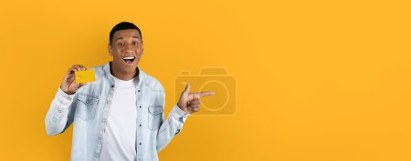 Téléchargez les photos : Homme joyeux montrant une carte de crédit et pointant avec enthousiasme vers un vaste espace vide sur un fond jaune de bannière web maquette - en image libre de droit