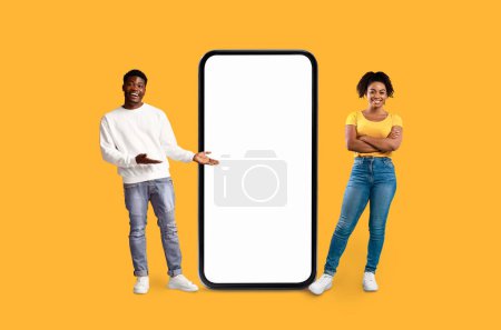 Foto de Sonriente hombre afroamericano que presenta algo con una mujer posando, ambos junto a una gran maqueta de teléfonos inteligentes contra una pared amarilla - Imagen libre de derechos