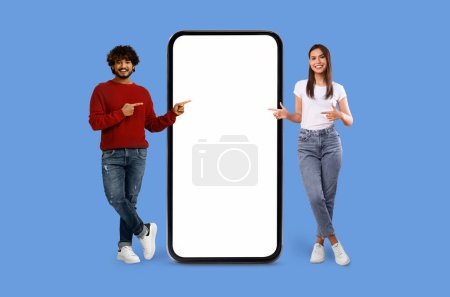 Téléchargez les photos : Joyeux jeune couple multiracial interagissant avec un grand écran de smartphone sur fond bleu, maquette - en image libre de droit