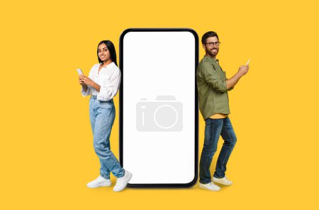 Téléchargez les photos : Un jeune homme et une jeune femme utilisant leur téléphone à côté d'un smartphone vierge maquette copier l'espace sur fond jaune - en image libre de droit