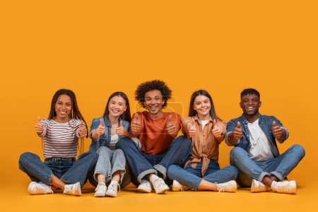 Eine enthusiastische multiethnische Gruppe junger Freunde, die Daumen nach oben streckt und Positivität und Übereinstimmung in einem modernen Umfeld repräsentiert, isoliert auf gelbem Hintergrund