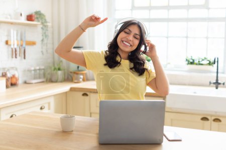 Foto de Una mujer árabe disfruta de la música en su computadora portátil con auriculares en la comodidad de su cocina casera, tienen descanso mientras trabajan - Imagen libre de derechos
