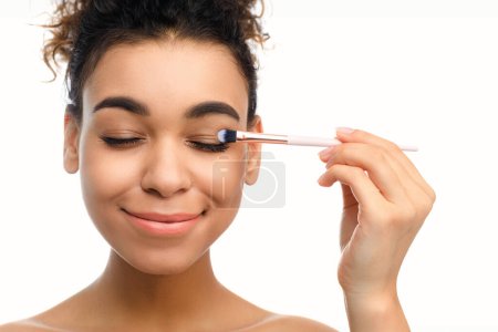 A black woman gently applies eye makeup using a brush, eyes closed, embodying a relaxed spa atmosphere and the beauty rituals of African American people
