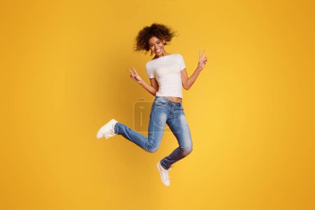 Foto de Chica milenaria afroamericana positiva saltando y mostrando gesto de paz, fondo naranja - Imagen libre de derechos