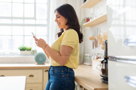 Foto de Ilustra una acogedora escena en casa, tal vez involucrando a una mujer de Oriente Medio, en una cocina iluminada por el sol con su teléfono inteligente - Imagen libre de derechos