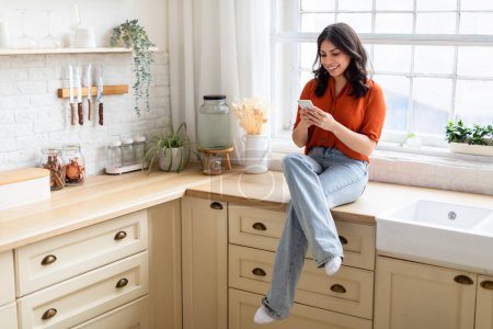 Téléchargez les photos : Une femme joyeuse du Moyen-Orient s'assoit sur un comptoir de cuisine, absorbée par son smartphone, copiant de l'espace - en image libre de droit
