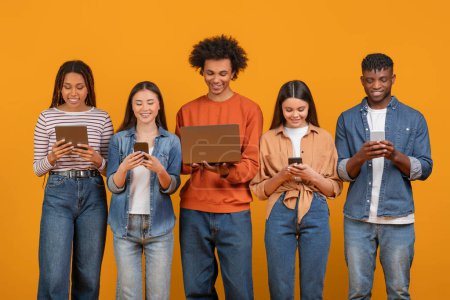 Photo for A focused international group of friends using various digital devices, showing the multiethnic, multiracial tech-savvy youth isolated against an orange background - Royalty Free Image