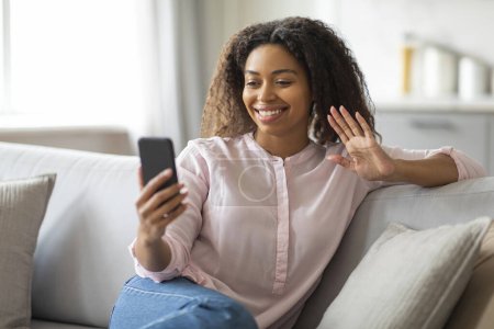 Photo for An african american millennial lady waves hello while video calling from her comfortable home, representing human connection - Royalty Free Image
