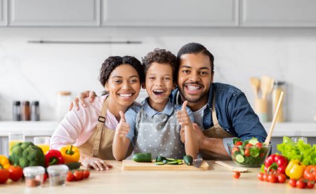 Foto de Familia afroamericana enérgica posando con los pulgares hacia arriba en la cocina, retratando un divertido momento de cocina familiar con comida saludable - Imagen libre de derechos