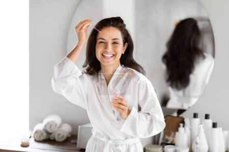A young woman in a white robe holding a skincare bottle smiles in front of a bathroom mirror, surrounded by toiletry items