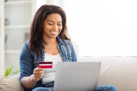 Foto de Mujer afroamericana está sentada en un sofá, sosteniendo una tarjeta de crédito en una mano y trabajando en una computadora portátil con la otra, enfocada y comprometida en una transacción en línea o actividad de compras.. - Imagen libre de derechos