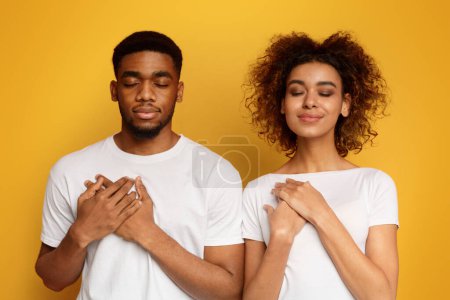 Grateful african-american couple keep both palms on chest with closed eyes, express gratitude and good feeling, orange background