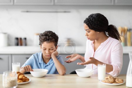 Téléchargez les photos : Cette scène représente une mère afro-américaine préoccupée s'adressant à son fils désintéressé pendant les repas à la table de la cuisine - en image libre de droit