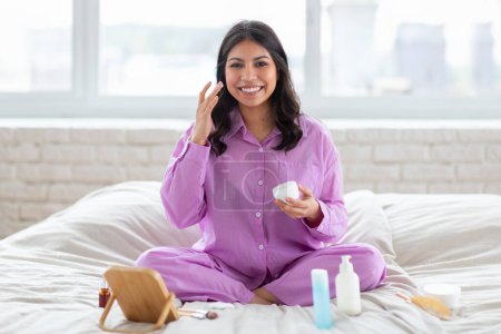 Photo for A cheerful arab woman enjoys a comfortable home setting, all while donning comfortable sleepwear, applying cream - Royalty Free Image