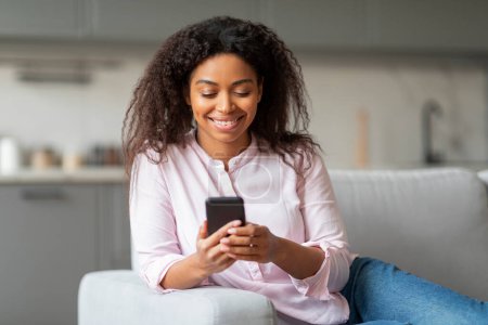 Téléchargez les photos : Une femme afro-américaine est vu confortablement textos sur son smartphone, offrant un aperçu de la vie à la maison et les habitudes millénaires - en image libre de droit