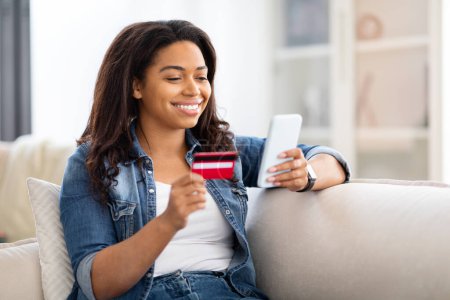Afroamerikanerin sitzt auf einer Couch, in der einen Hand eine Kreditkarte, in der anderen ein Handy. Sie scheint sich auf den Telefonbildschirm zu konzentrieren, während sie ihre Finanzen verwaltet.