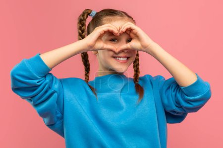 Cute teen girl showing heart gesture over her face and smiling, posing isolated on pink studio background