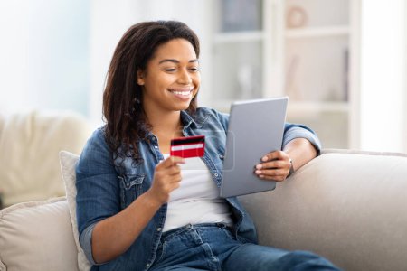 Téléchargez les photos : Femme afro-américaine est assise sur un canapé, tenant une carte de crédit dans une main et une tablette dans l'autre, concentrée sur l'écran, effectuant éventuellement un achat en ligne ou gérant ses finances. - en image libre de droit